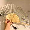 A hand is holding a light-colored folding fan with black accents and leaf patterns.