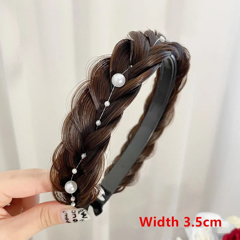 A hand holds a black hair band featuring dark brown braided hair and small pearl embellishments.