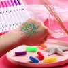 A hand is painting a flower design on skin using colorful dual-ended watercolor art markers placed on a pink tray.