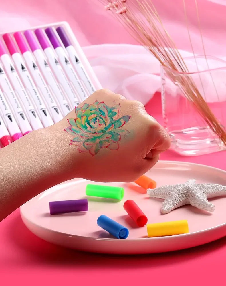 A hand is painting a flower design on skin using colorful dual-ended watercolor art markers placed on a pink tray.