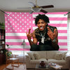 A man with dreadlocks wearing a dark shirt sits in front of a pink and white striped flag banner.