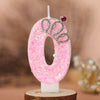 A pink glitter number zero candle decorated with a silver crown and a red gemstone, set against a blurred background.