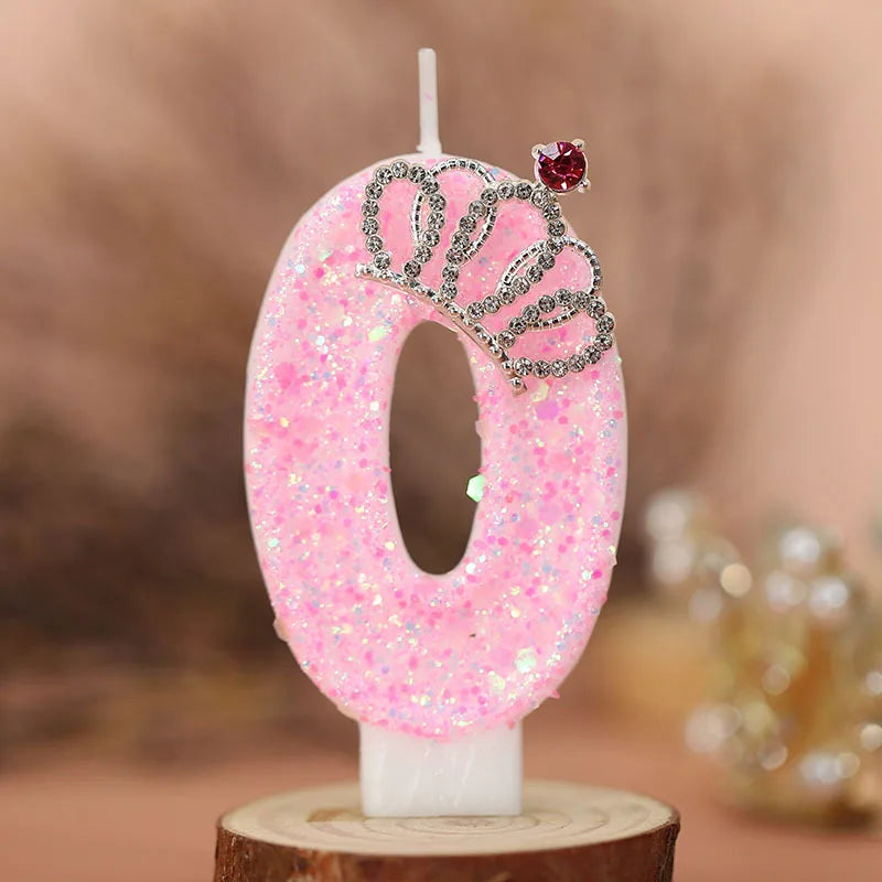 A pink glitter number zero candle decorated with a silver crown and a red gemstone, set against a blurred background.