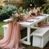 A rustic wedding setup featuring a white table draped with pink tulle fabric and floral arrangements in an outdoor garden set