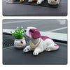 A white and pink dog ornament figure is shown resting on a dark gray surface next to a small potted plant.