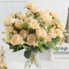 A bouquet of light peach and cream artificial roses with green foliage displayed in a glass vase.
