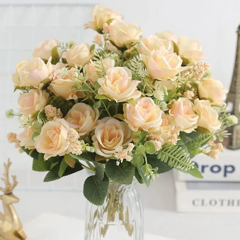 A bouquet of light peach and cream artificial roses with green foliage displayed in a glass vase.