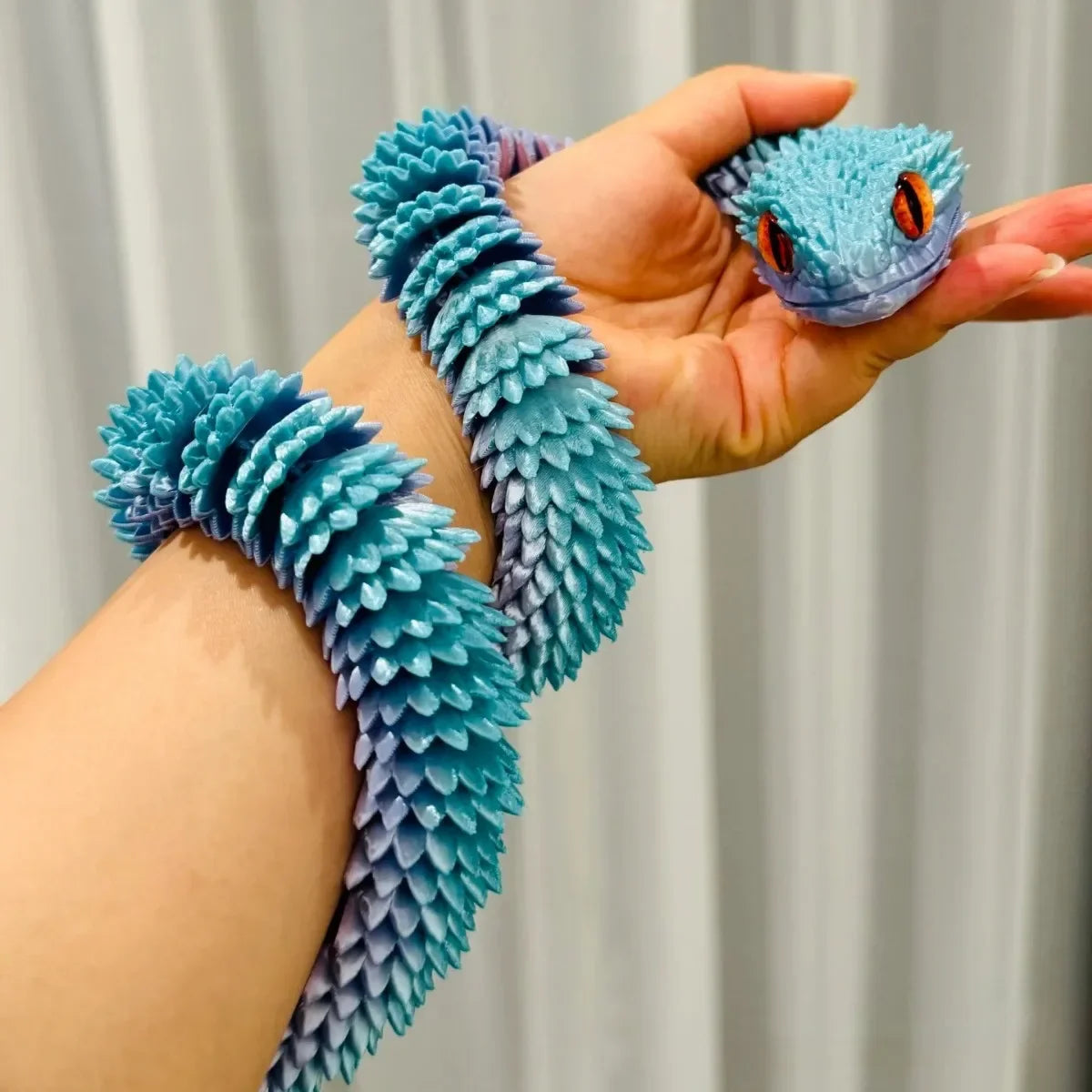 A hand holds a blue and purple 3D printed articulated snake fidget toy with orange eyes.