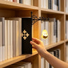 A hand reaches for a black book with an ornate iron hanger lamp on a wooden bookshelf.