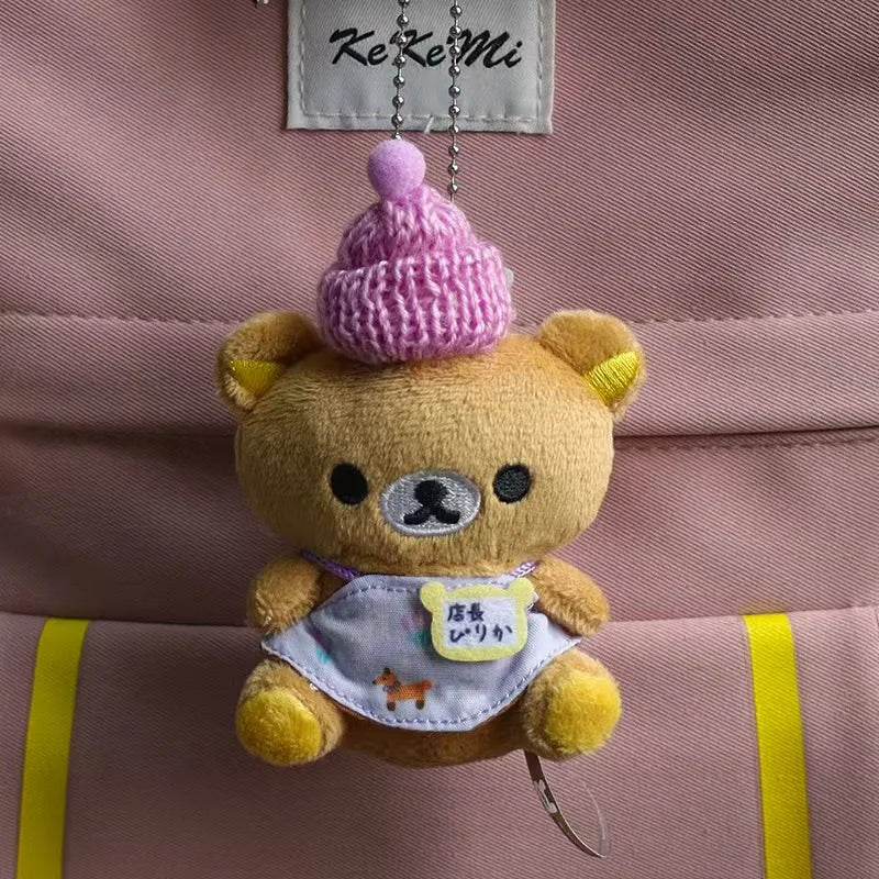 A fluffy brown Rilakkuma plush bear with a pink hat and a white bib is attached to a pink fabric bag.
