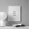A framed motivational quote print on a gray wall with an open book and metallic objects on a white surface.