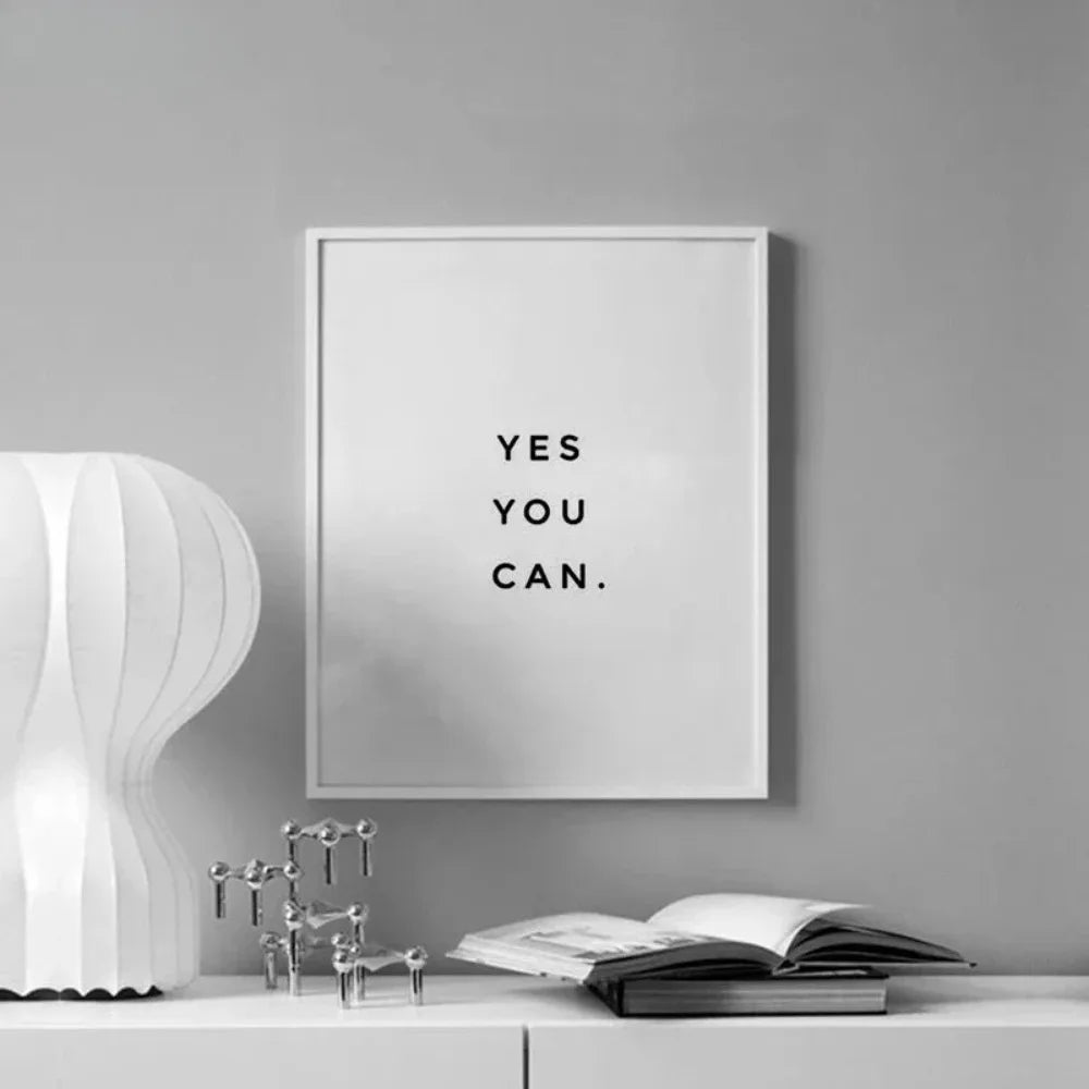 A framed motivational quote print on a gray wall with an open book and metallic objects on a white surface.