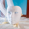 A white, sculpted masquerade mask with intricate tree-like designs is displayed on a light fabric surface.