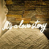 A white knitted fabric rests in front of a rustic stone wall background with a glowing neon sign that reads Its a love story