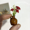 A small brown ceramic vase holds artificial red flowers, being held by a hand.