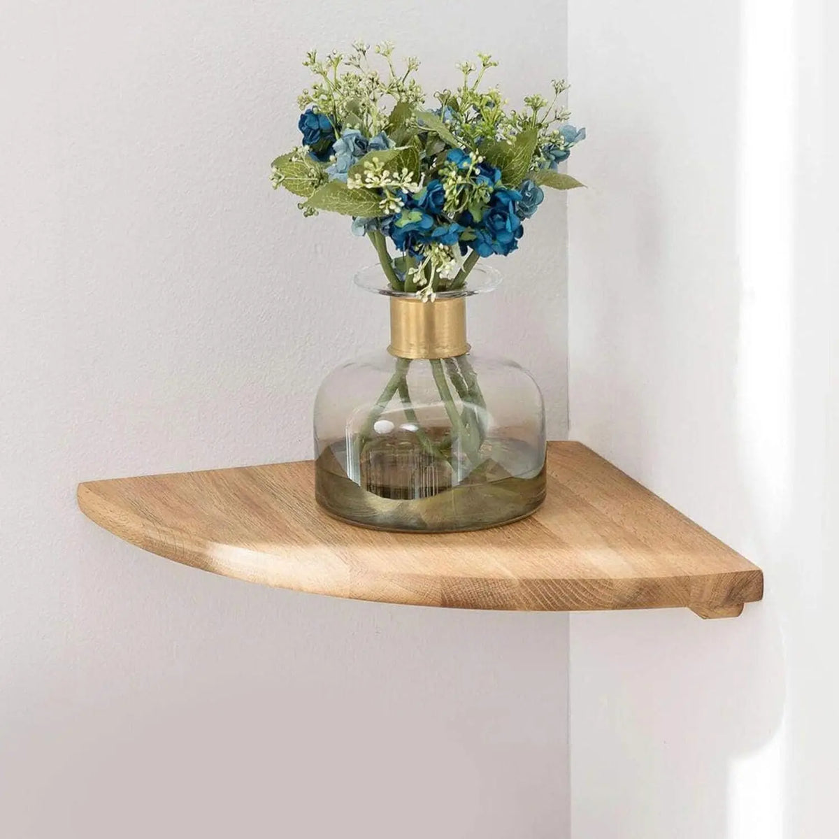 A glass vase filled with blue and green flowers rests on a natural wooden corner floating shelf.