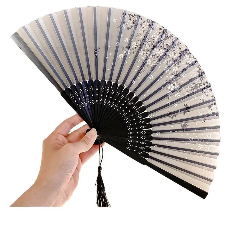 A hand holds an elegant floral bird print bamboo folding fan, showing its pleated design and light beige color.