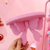 A hand holds a pink decorative bow-shaped wooden wall shelf against a light pink background.