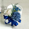 A bouquet of dusty blue silk roses and blue flowers tied with a deep blue ribbon in a white cup.
