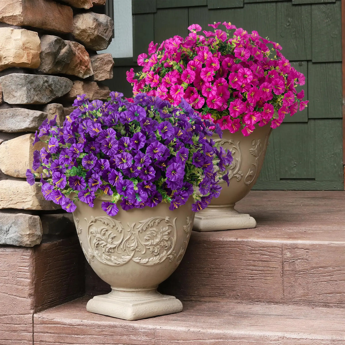 A display of vibrant purple and magenta artificial silk flowers in decorative urns set against a stone wall and wooden steps.