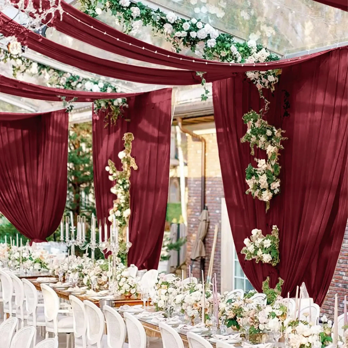 Deep burgundy sheer chiffon curtains draped with white floral garlands for a wedding event backdrop.