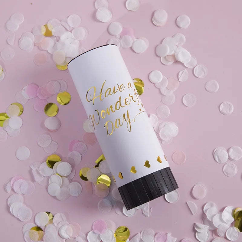 A white hand twist confetti cannon is shown surrounded by pink and gold confetti petals on a light pink background.