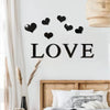 Black acrylic heart wall stickers spelling the word LOVE displayed on a white wall above a wooden bed frame.