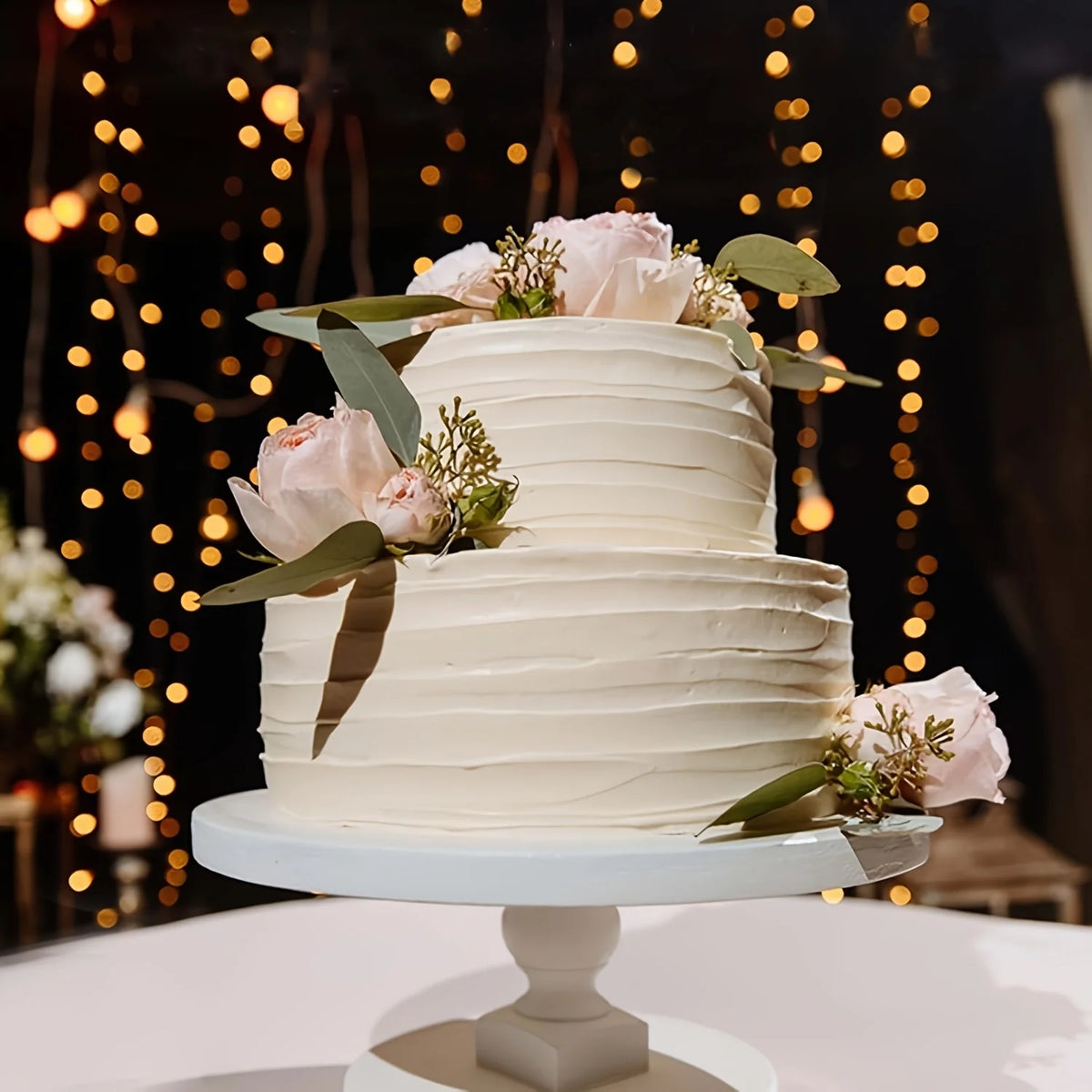 A two-tiered white cake decorated with pink roses and green leaves, displayed on a stand against a dark background with warm 