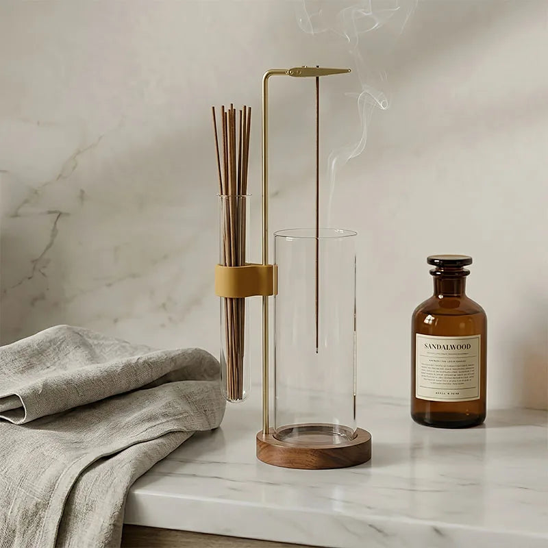 Un haut cendrier en verre avec support en laiton et bâtons en bois, posé sur une surface de marbre à côté d'une bouteille en 