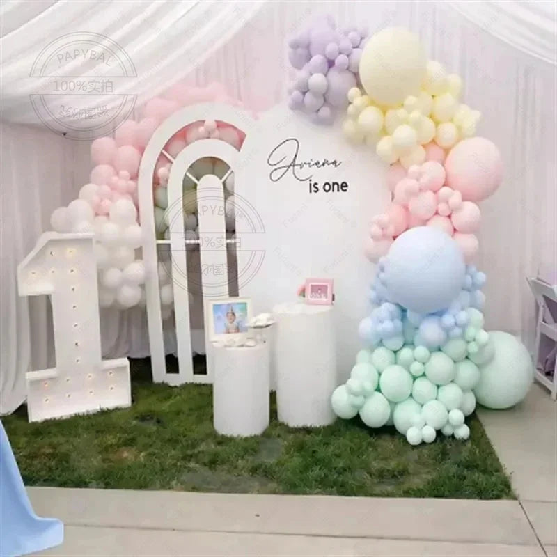 A pastel balloon garland arch kit featuring assorted rainbow colors and white balloons for a first birthday celebration backd