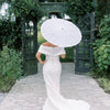 A woman in a white dress holds a large white paper parasol under an arched trellis in a garden setting.