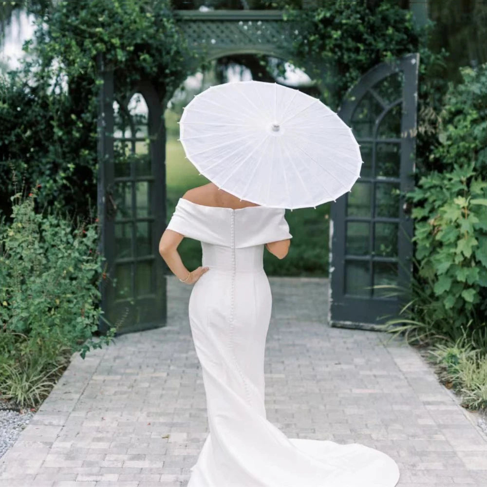 A woman in a white dress holds a large white paper parasol under an arched trellis in a garden setting.