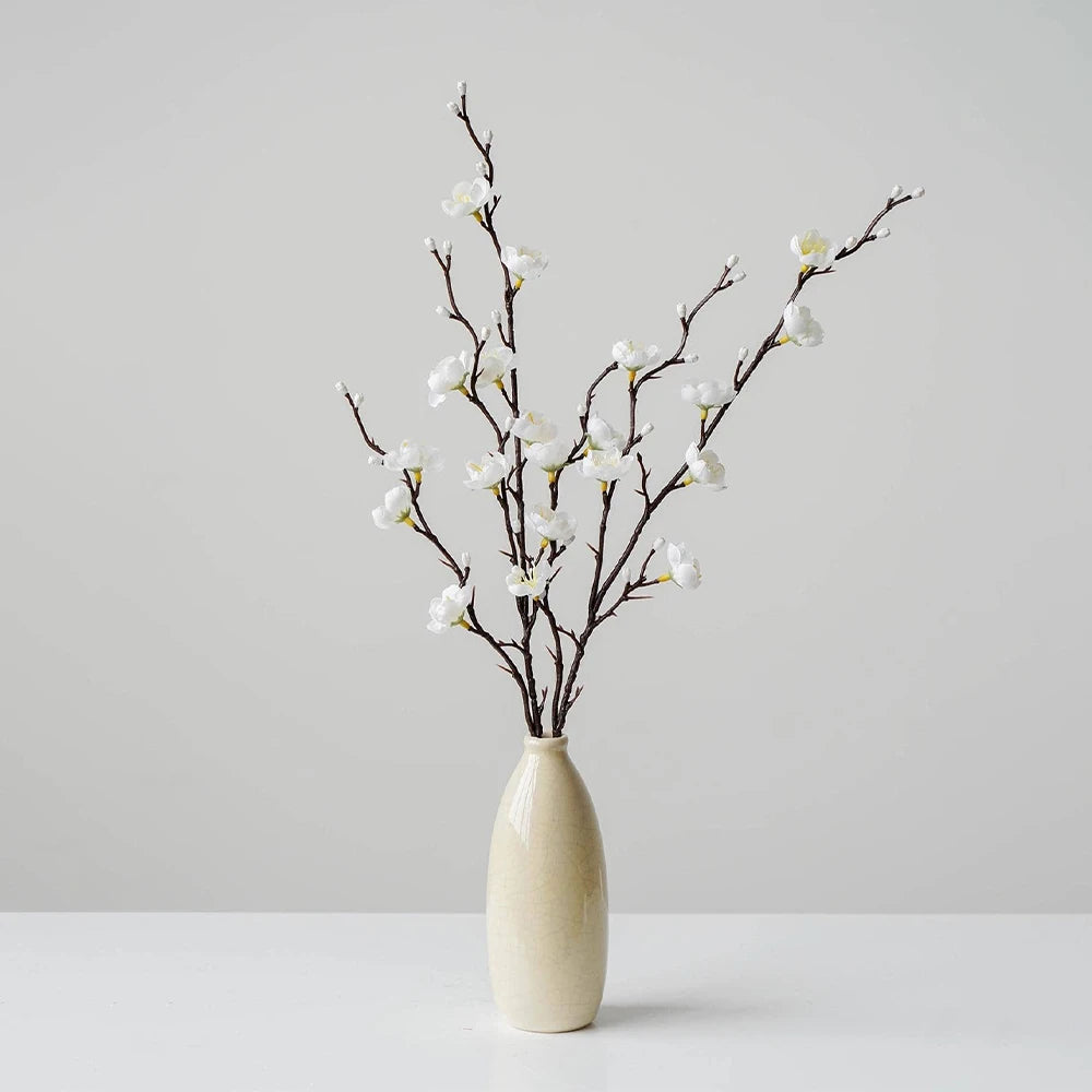 A vase holds delicate artificial plum blossom branches with white flowers against a light gray background.