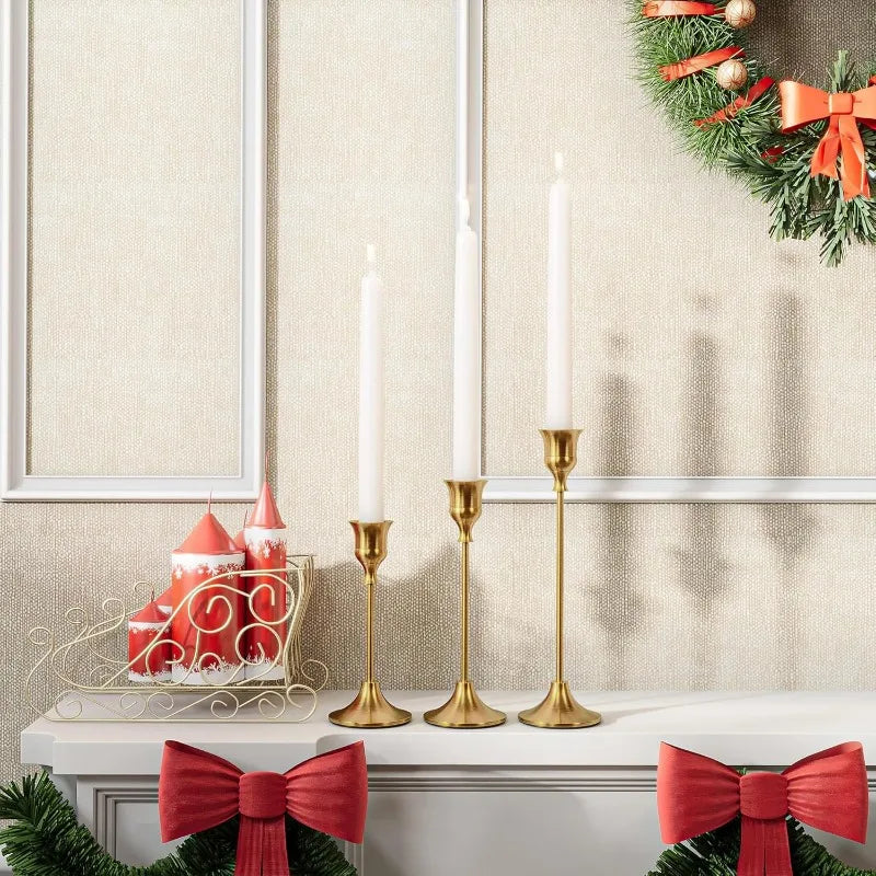 A set of three gold-toned metal candle holders with white taper candles displayed on a white mantelpiece.