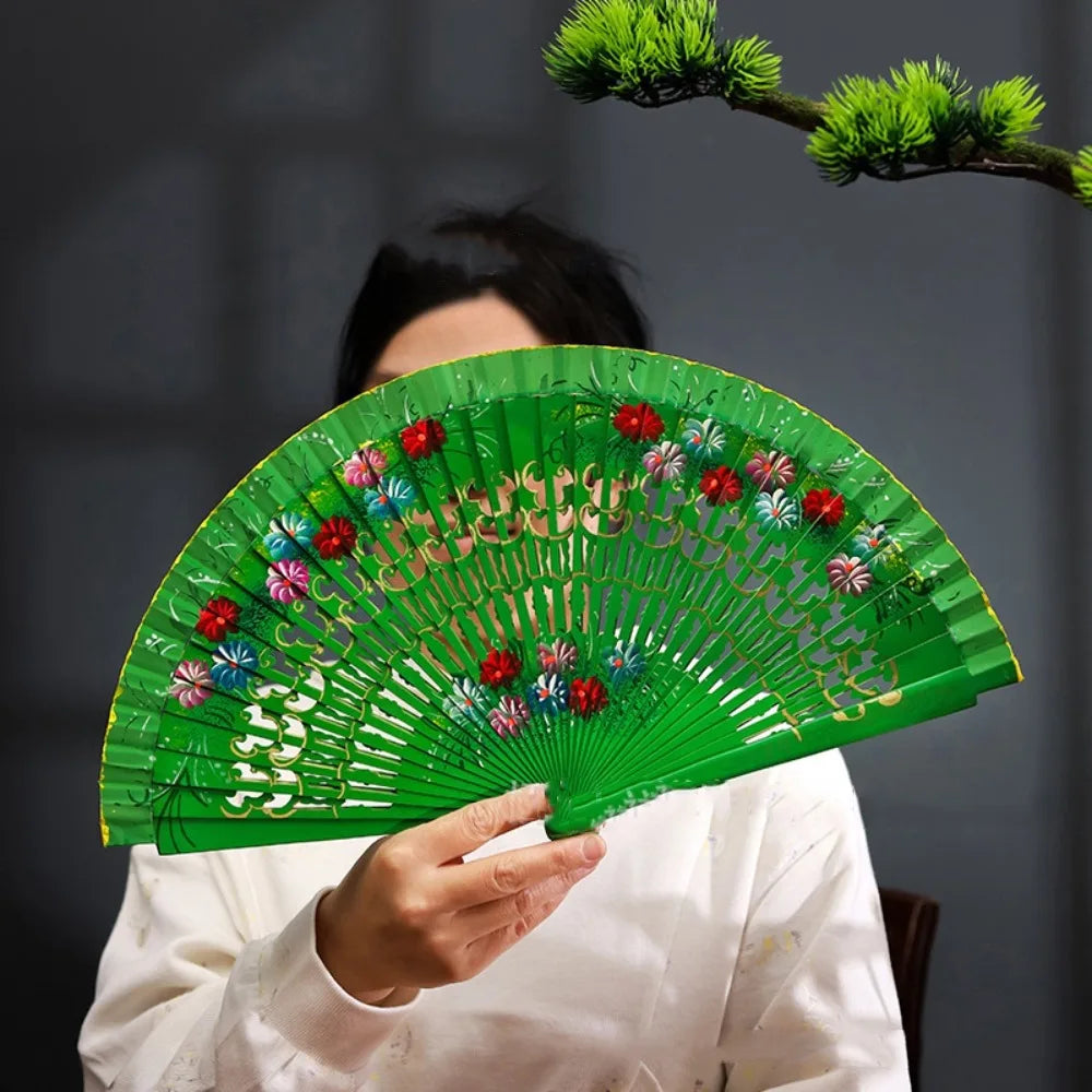 A hand-painted decorative folding fan with green slats and floral accents is held by a person against a dark background.