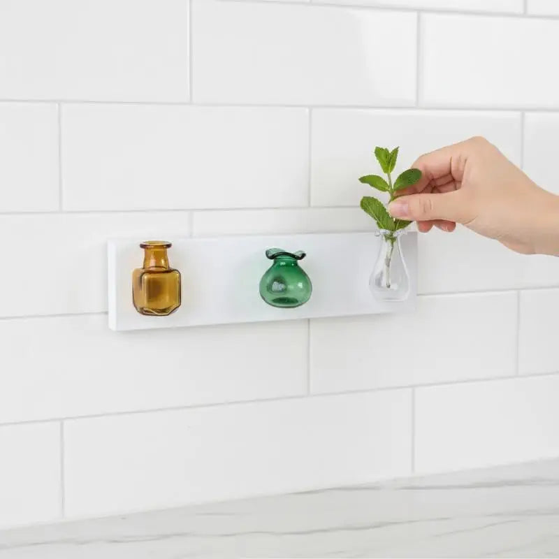 A hand places a small glass flower vase onto a magnetic white tile on a kitchen backsplash.