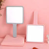 A pink compact handheld makeup mirror with a silver frame and ergonomic handle displayed on a pink surface.
