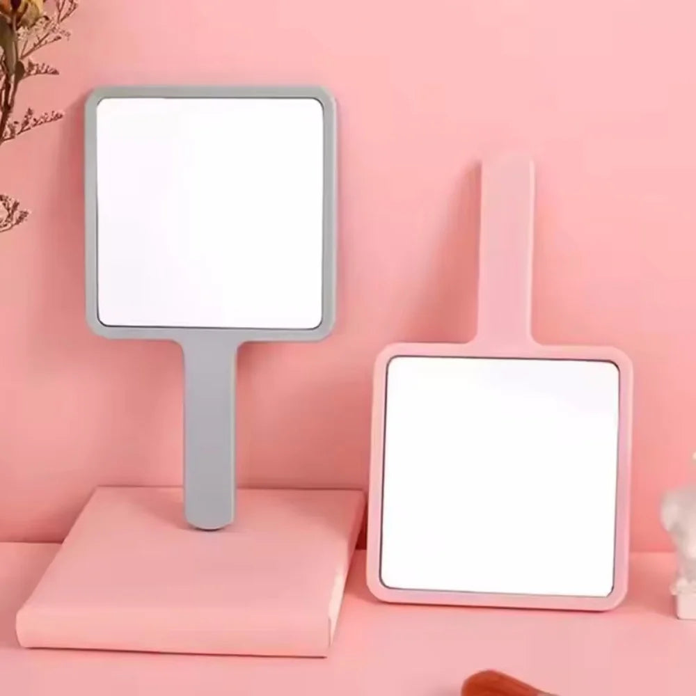 A pink compact handheld makeup mirror with a silver frame and ergonomic handle displayed on a pink surface.