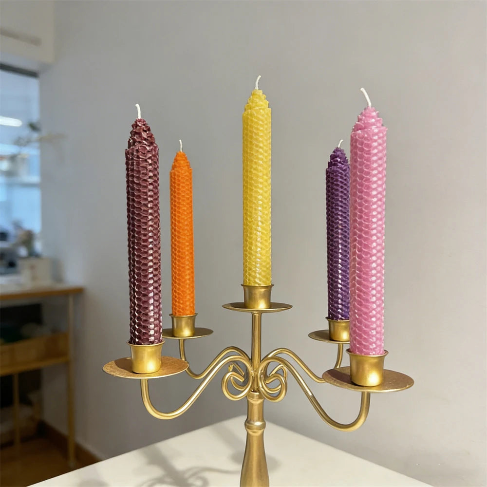 A display of six colorful honeycomb beeswax candles in various shades of purple, orange, and yellow on a brass candle holder.