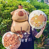 A person holds a large foil balloon shaped like a capybara bear and another round capybara balloon in an outdoor setting.