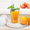 A glass of orange juice garnished with mint on a woven coaster next to a whole orange.