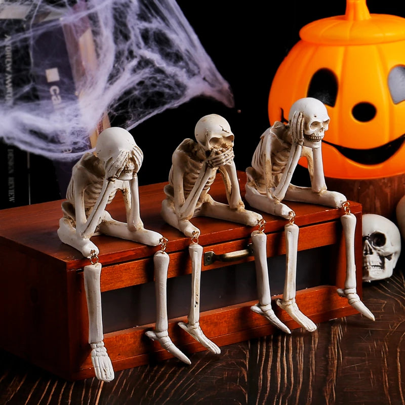 A set of three white resin skeleton skull figurines are displayed on a wooden box next to a carved orange jack-o'-lantern.