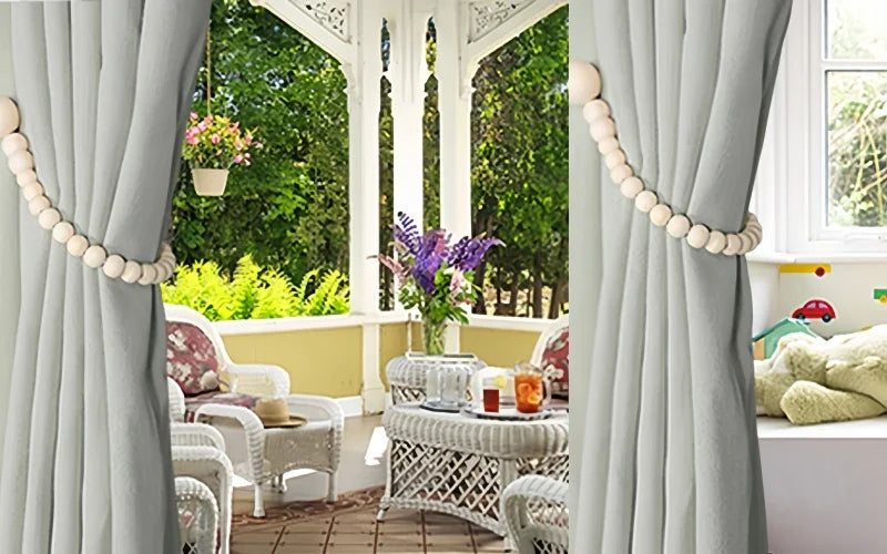 Gray fabric curtains with wooden bead tiebacks framing a garden view and antique furniture.