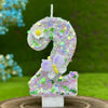 A large purple glitter number 2 cake candle decorated with a white flower and a light purple butterfly, set against a green o
