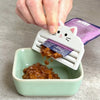 A hand is squeezing cat food from a purple bag into a green bowl using a white plastic squeezer tool.