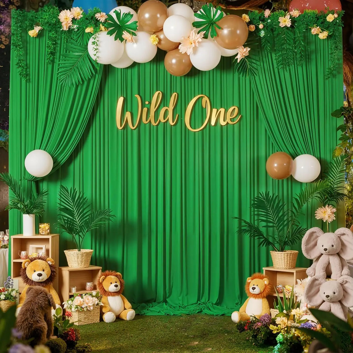 A green fabric backdrop decorated with gold lettering Wild One and floral greenery and balloons for a first birthday party