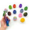 A hand holds a variety of colorful, textured 3D printed dragon egg fidget toys scattered on a white surface.