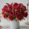 A vibrant bouquet of deep red and burgundy artificial silk flowers arranged in a white textured vase.