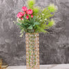 A tall crystal gold vase filled with pink roses and green foliage on a white marble surface.