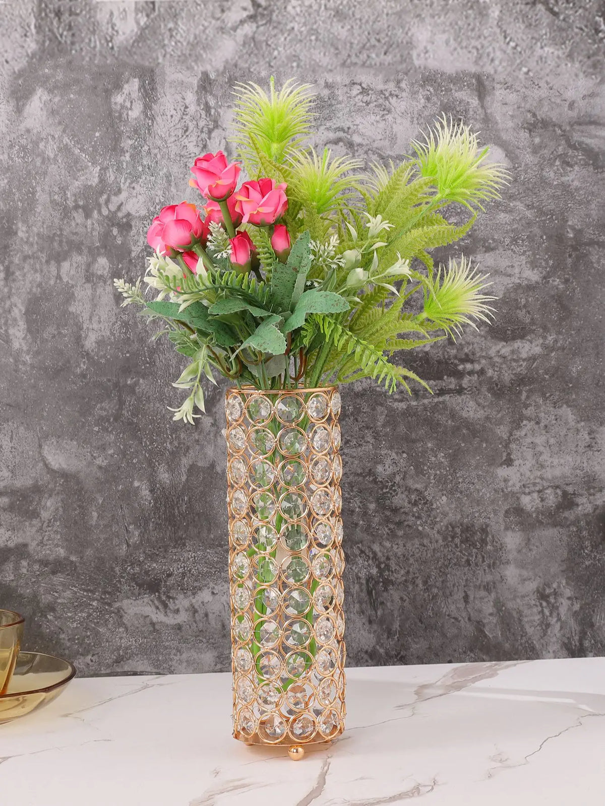 A tall crystal gold vase filled with pink roses and green foliage on a white marble surface.